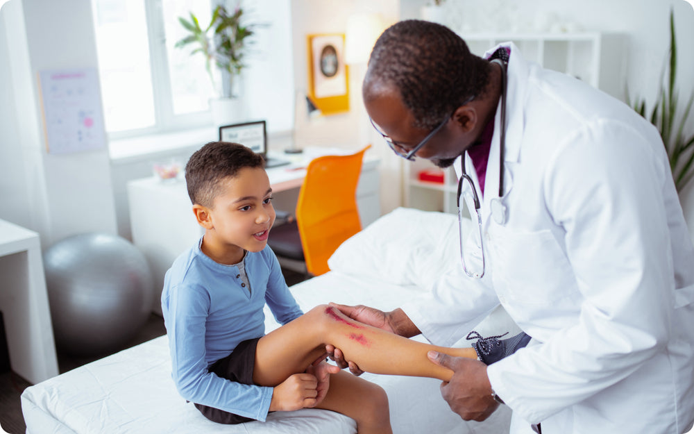 Doctor in lab coat and stethoscope looking at scrape on child patient's knee/shin area. 